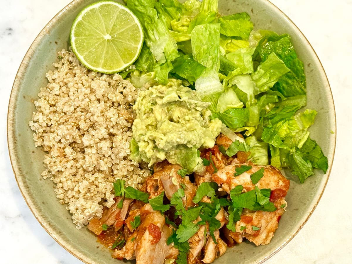 slow cooker mexican chicken in a blow with quinoa, guacamole and garnished with lime and chopped cilantro