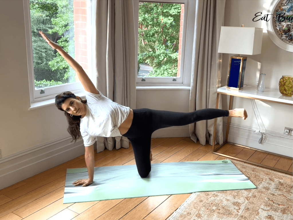 Woman on yoga mat on one knee with one hand on the floor the other in the air and a leg lifted to the side